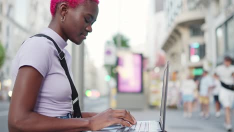 young woman with pink hair working on a laptop on a busy city street