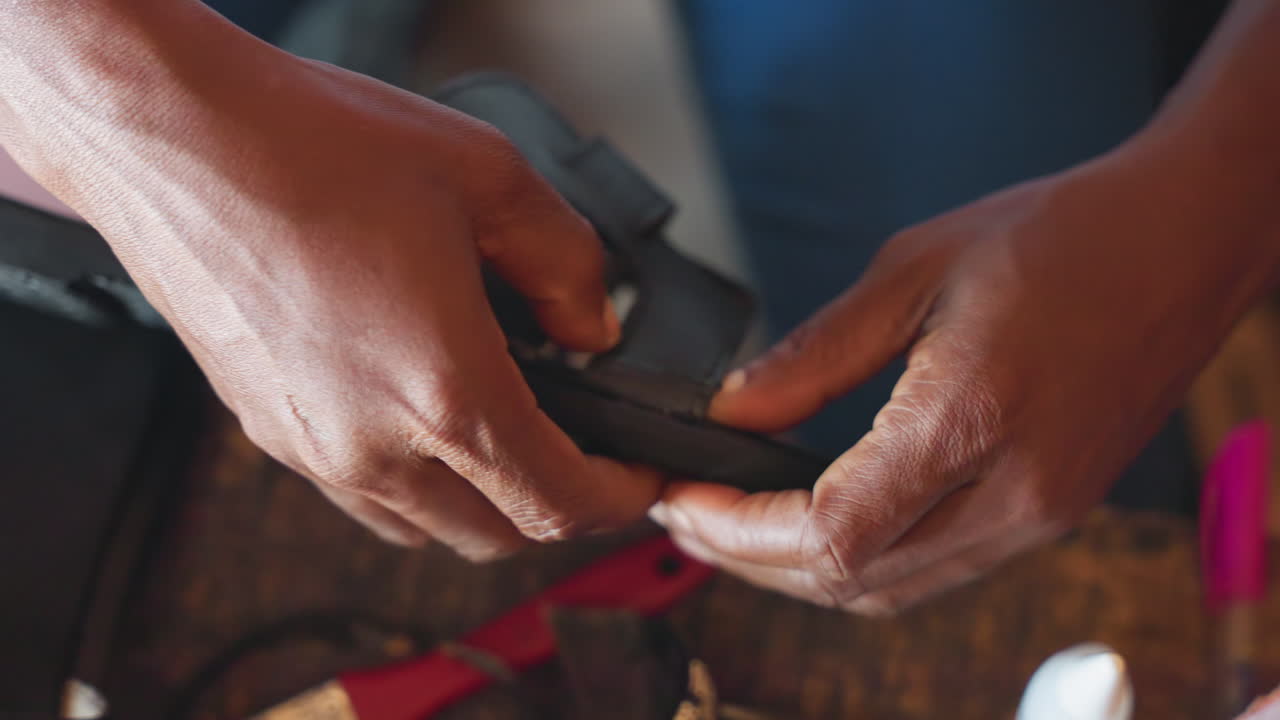Tangan memasang tali selama proses perbaikan sepatu. Pengrajin dengan cermat memasukkan gesper melalui lubang kulit saat bekerja. Pemandangan pedesaan perbaikan sepatu dengan alat-alat yang tersebar dan pengerjaan kulit yang detail.