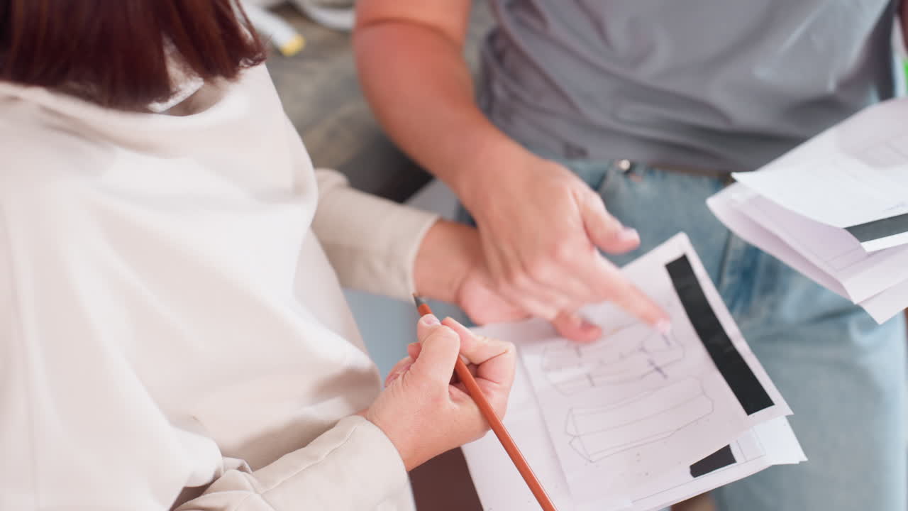 Fashion designer explaining trouser design to assistant while pointing at sketch on white paper during creative discussion session focusing on planning garment details and clothing development
