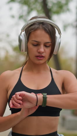 woman in black singlet and headphones checking time on wristwatch in park, surrounded by trees, maintaining focus during workout session while keeping track of time for her exercise routine