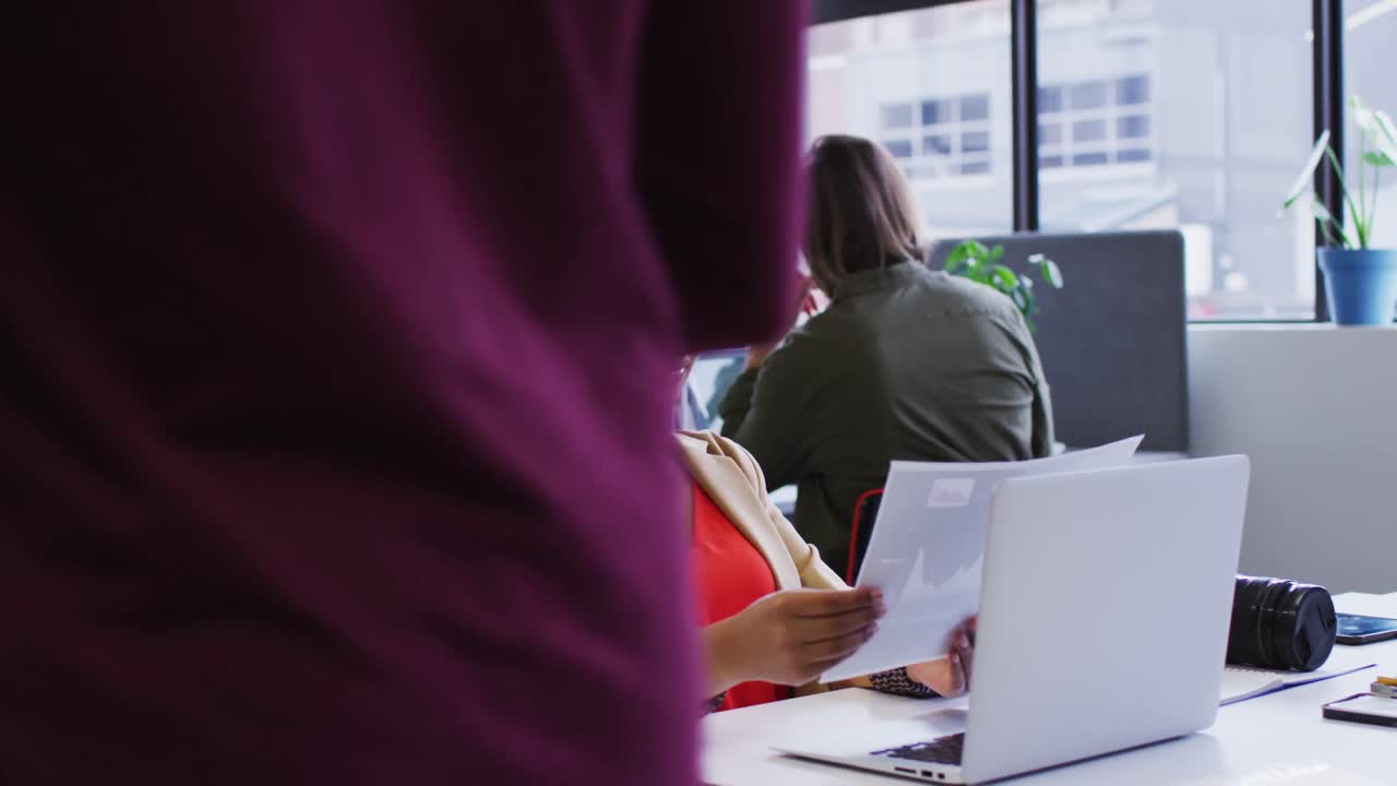 mujer de negocios de raza mixta sentada usando una computadora portátil pasando por el papeleo en una oficina moderna