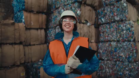 una chica morena en un casco blanco un uniforme azul y un chaleco protector naranja sostiene una tableta en sus manos se para y mira a la cámara cerca de una enorme pila de almacenamiento de residuos reciclados