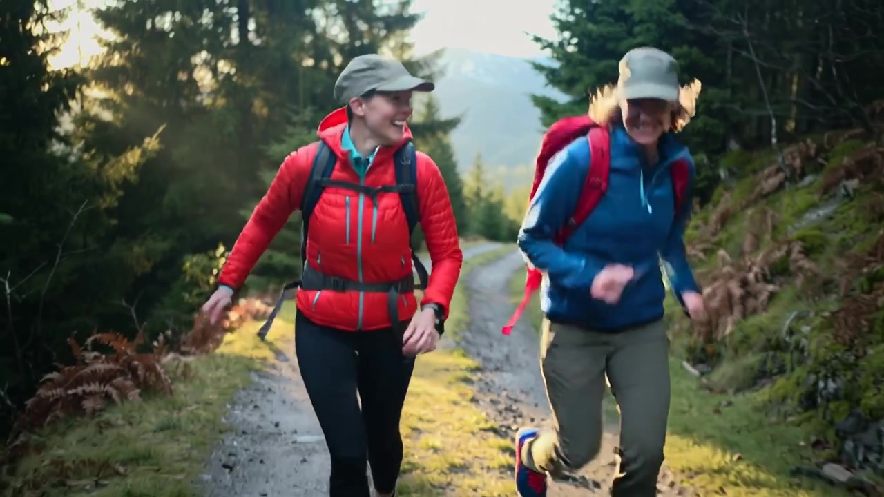 Due escursionisti che camminano ridendo, scambiandosi sguardi, aumentando il passo fino a fare jogging su un sentiero nel bosco con zaini da giorno e cappellini.