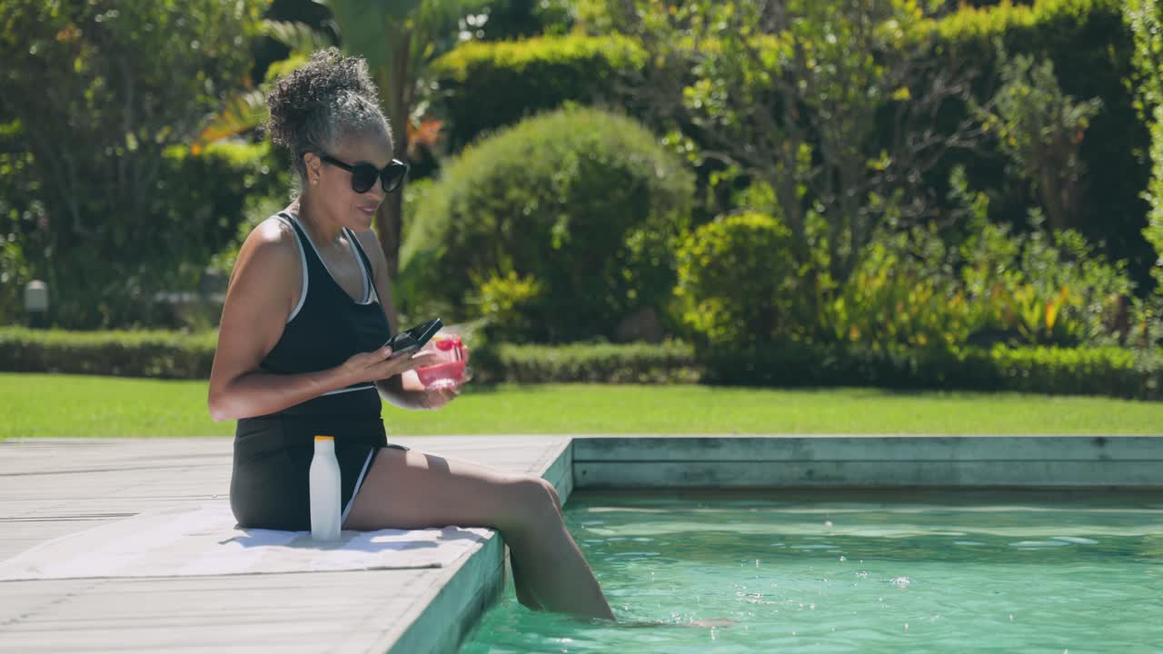 Afroamerikanische Frau im Badeanzug am Pool, die ein pinkes Getränk trinkt, am Telefon Nachrichten checkt und ein Selfie macht