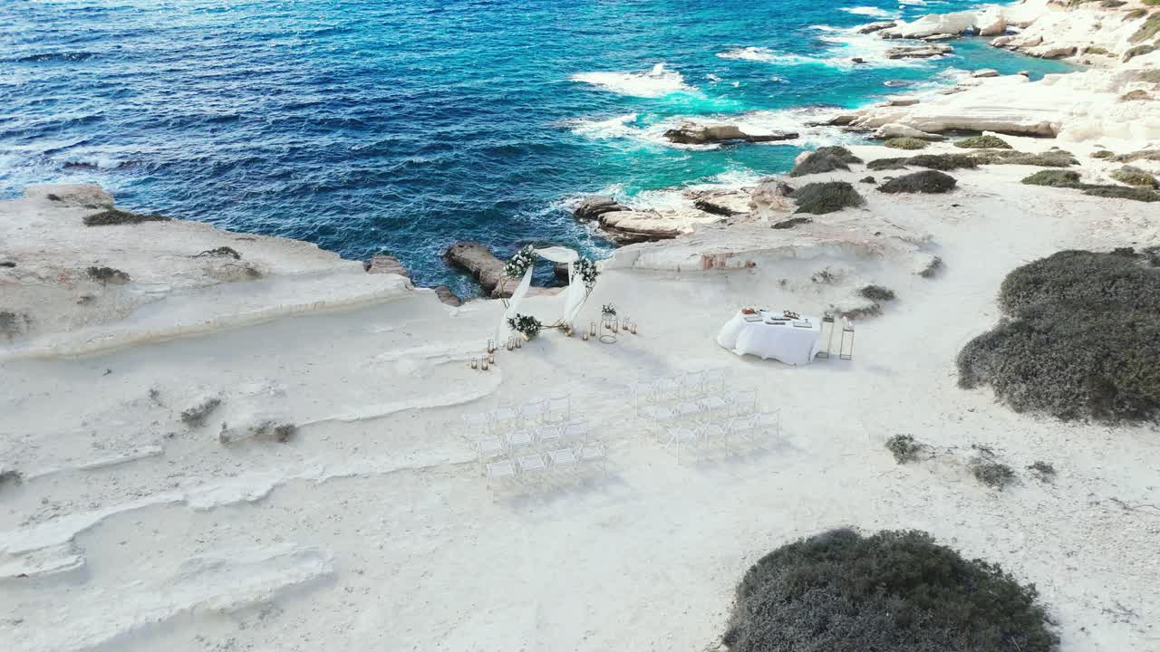Stunning wedding setup by the sea features elegant chairs and a decorated altar against a backdrop of clear blue water and rocky terrain. The scene is perfect for a romantic ceremony