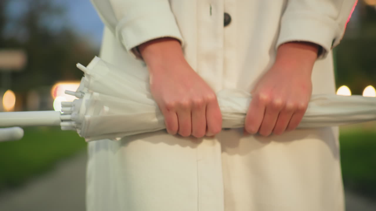 Close up of person in white coat firmly squeezing folded umbrella with both hands outdoors, soft bokeh lights of moving cars and green background create calm evening urban atmosphere
