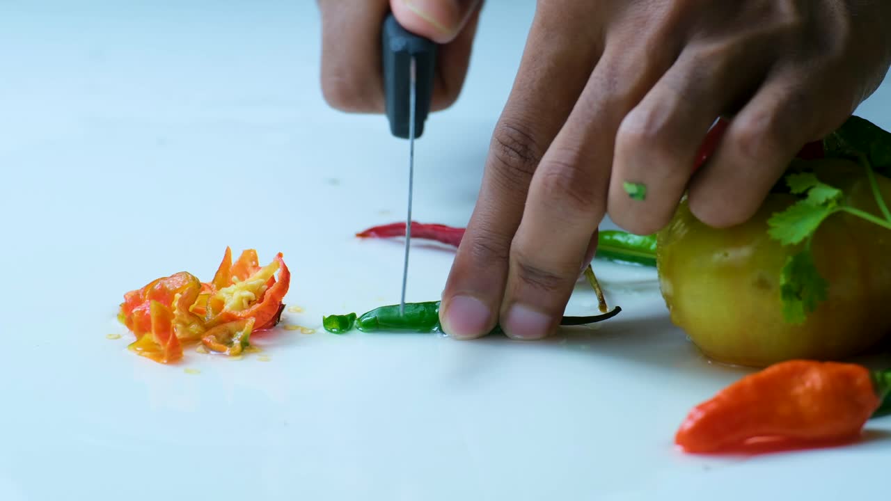 cerca del clip de las manos cortando finamente el chile verde con un cuchillo de cocina, al lado de otros vegetales y fondo blanco