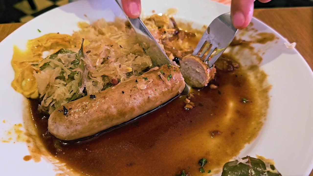 Beyaz bir tabakta lahana turşusu, hardal ve kahverengi sos ile Alman bratwurst kesen bir kişinin yakın çekim videosu, geleneksel Bavyera konfor yemeği ile sıcak bir restoran yemek sahnesi