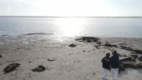 aerial-of-an-older-couple-with-mans-arm-around-the-woman-looking-out-into-the-sunset-on-the-beach