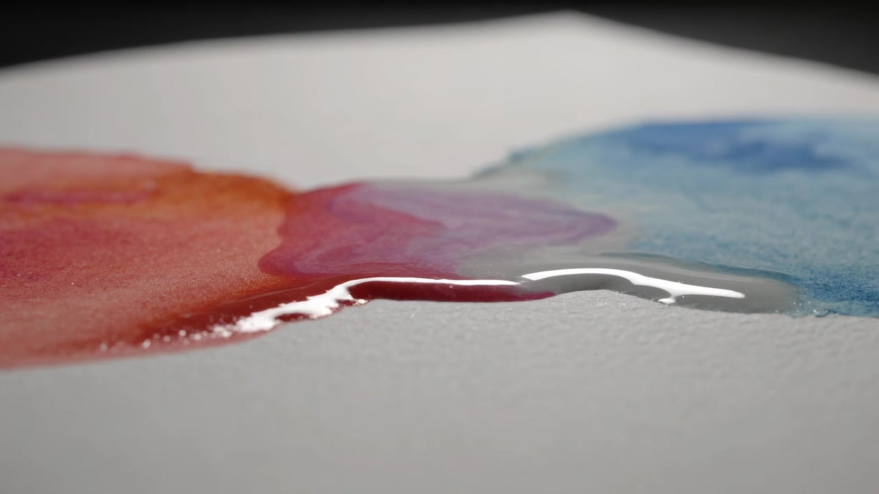 Una pellicola d'acqua che si espande, trascinando pozzanghere rosse e blu su carta acquerello testurizzata, formando il colore viola.