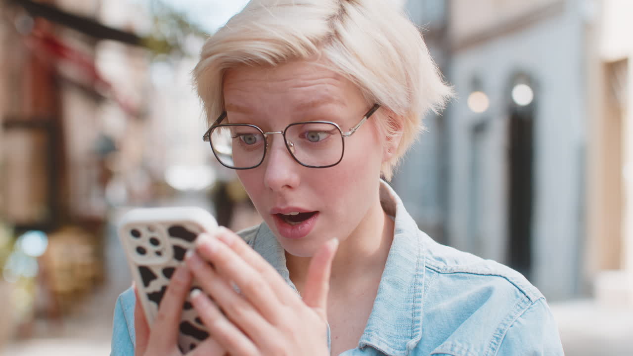 Ung kvinde reagerer med chok og overraskelse, mens hun kigger på sin smartphone på en bygade.
