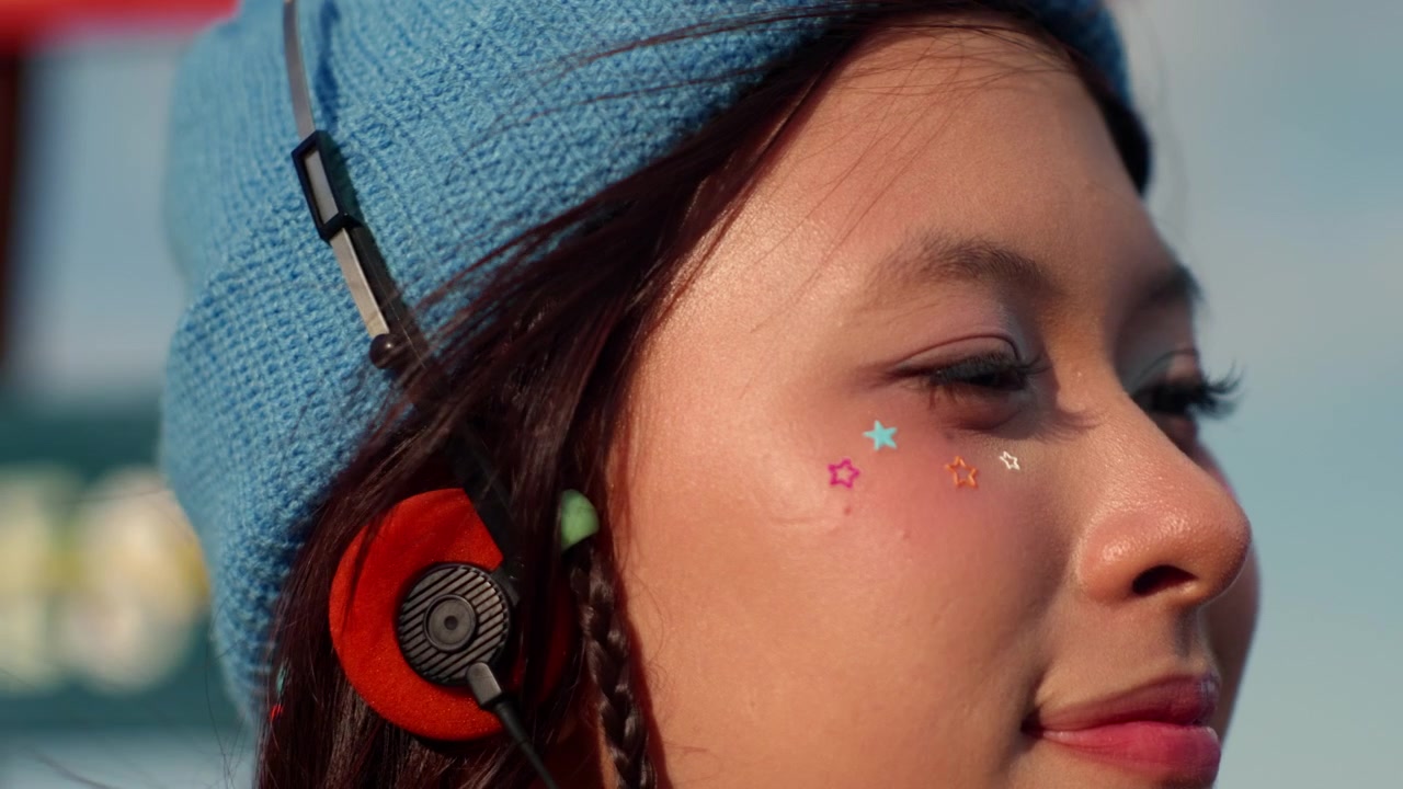Giovane donna asiatica con stelle glitterate sul viso indossa cuffie arancioni e un berretto blu in un ritratto ravvicinato illuminato dal sole, con un sorriso dolce e felice.