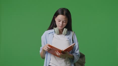 asian teen girl student with a backpack reading book and yawning while standing in the green screen background studio