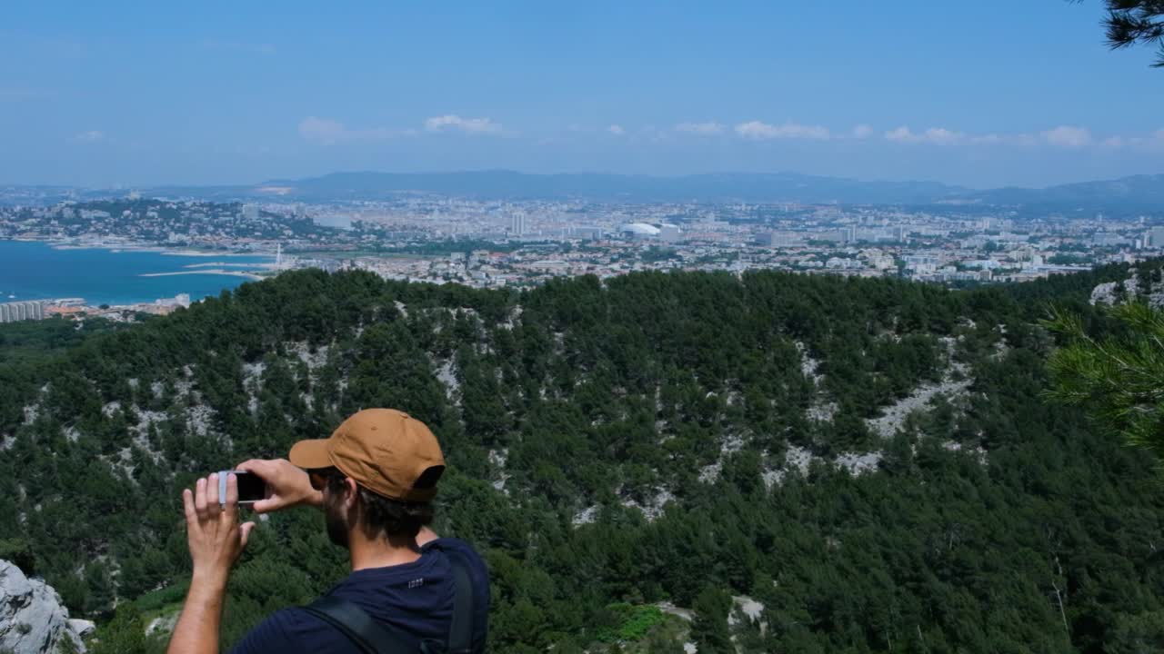 Man taking a picture with his phone from the top of Marseilleveyre, the mountain dominating the city of Marseille in the south of France on the mediterranean coast. Scenic view of Marseille and sea.