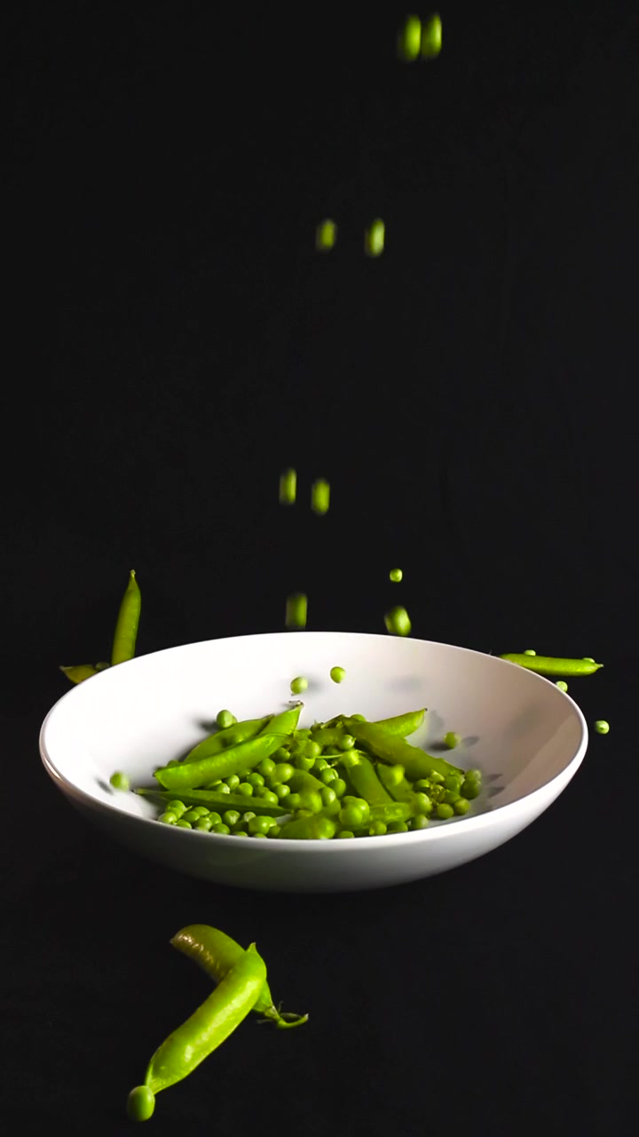 Fresh green peas falling into a white bowl full of pods on a black background, representing a healthy diet with natural ingredients. Capturing the essence of vegetarian food and cooking
