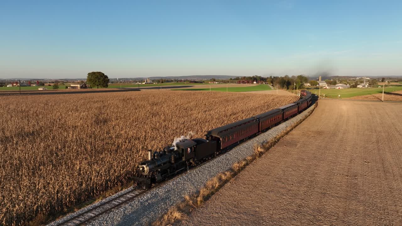 A steam train moves along railway tracks surrounded by vast cornfields as the sun sets, casting a warm, golden glow over the scenic rural landscape and nearby farms.