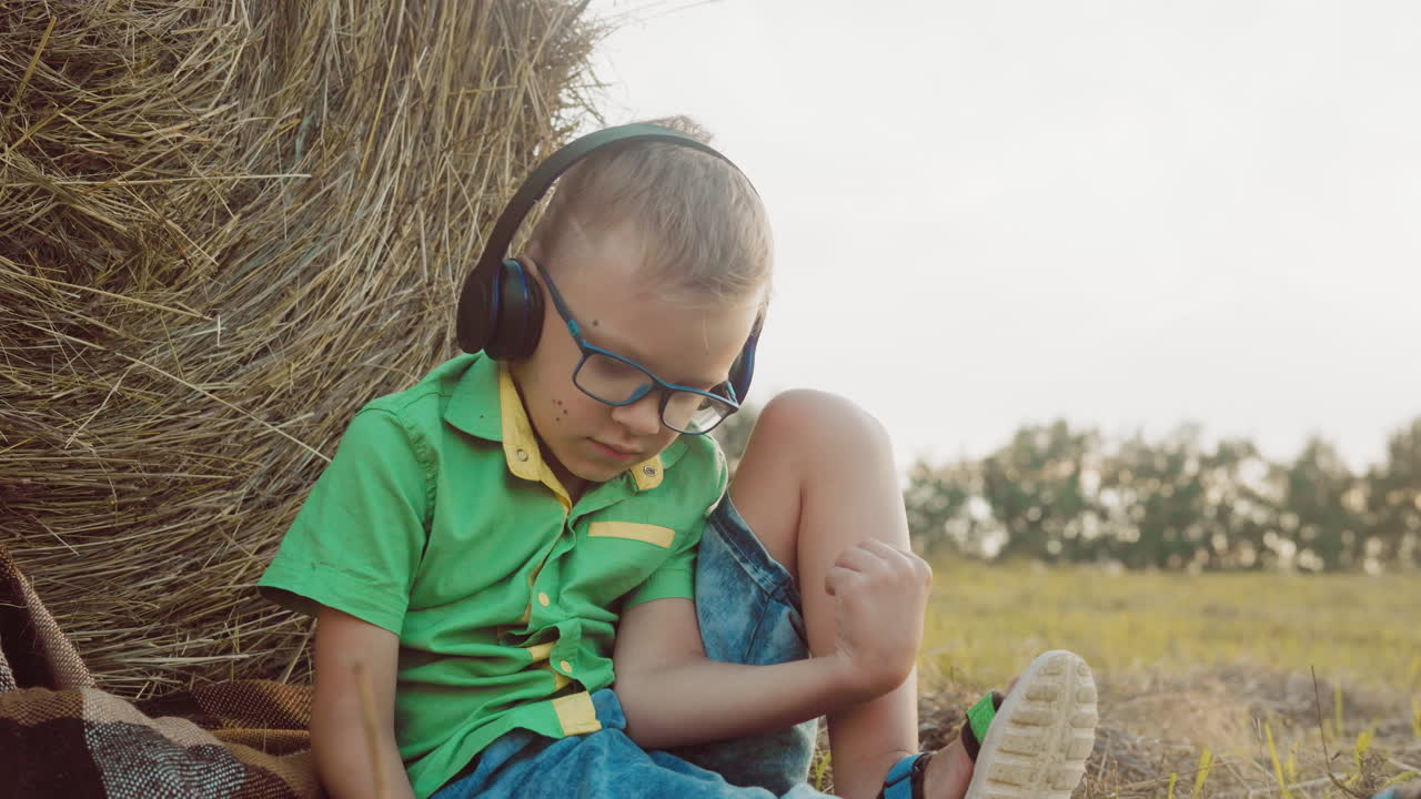 niño pequeño con auriculares profundamente inmerso en la música, sentado en una manta con la cabeza inclinada hacia abajo, rodeado de balas de heno en un entorno rural sereno durante la puesta del sol
