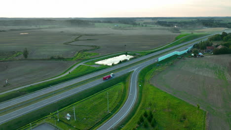 aerial shot of highway cutting through vast farmland with vehicles, featuring a small pond and rural scenery under soft evening light