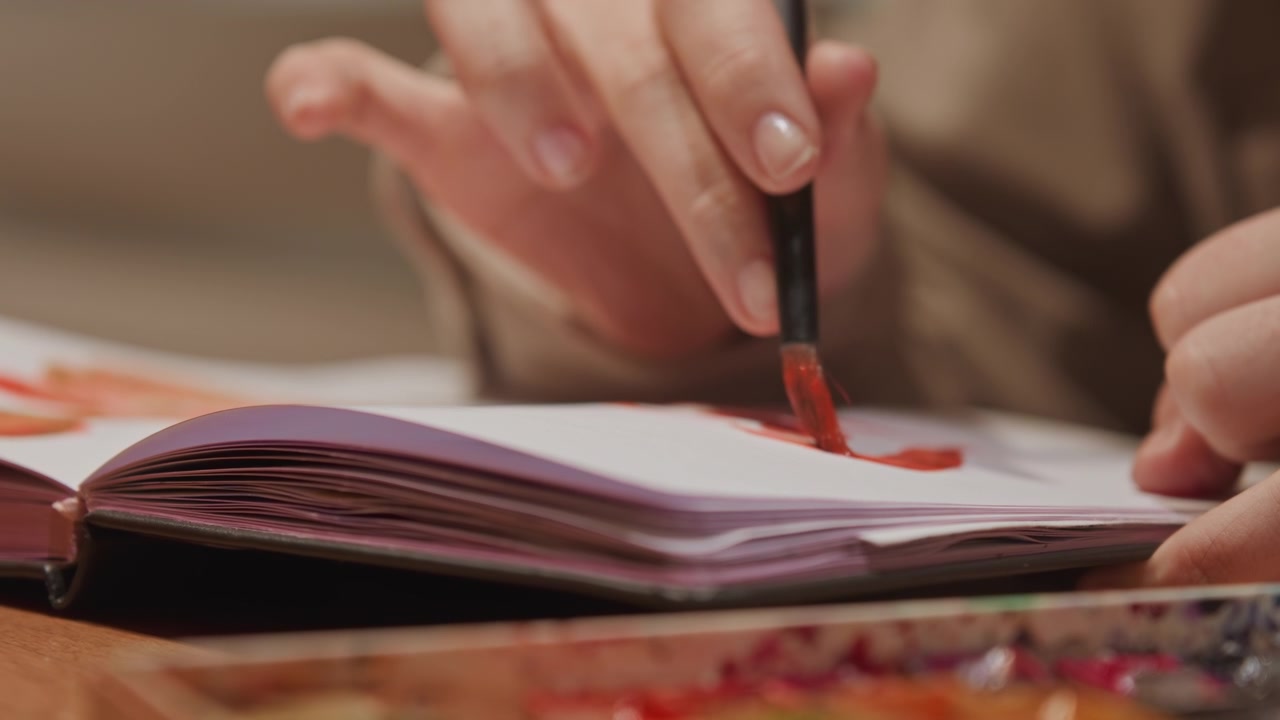 Close up of person hands painting with red watercolor on paper carefully applying paint to create artwork. scene shows texture of thick white pages in open sketchbook and various colors