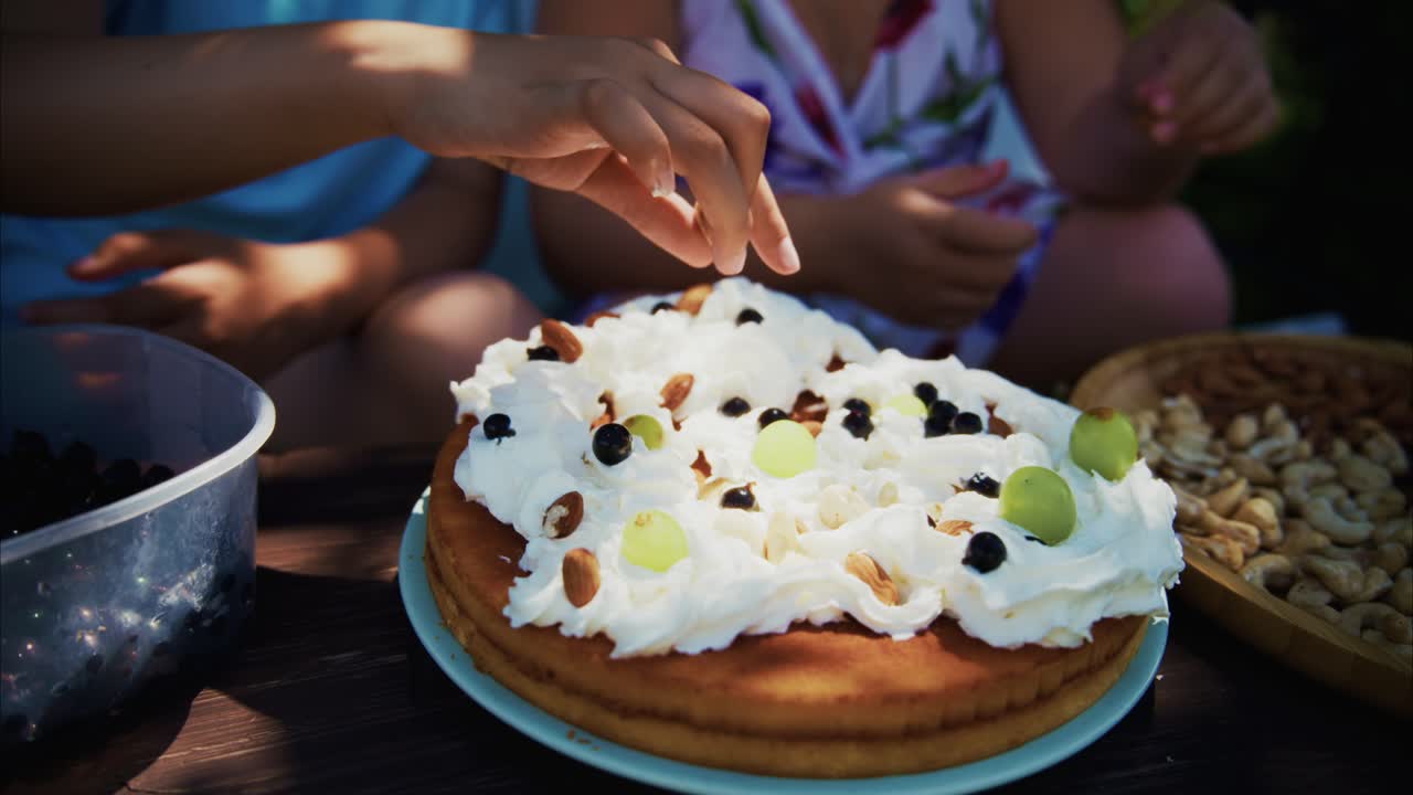 Hand placing toppings on a delicious cake with whipped cream and fresh fruits in a sunny outdoor setting