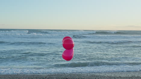 three red balloons float in wind as sea waves break in the background