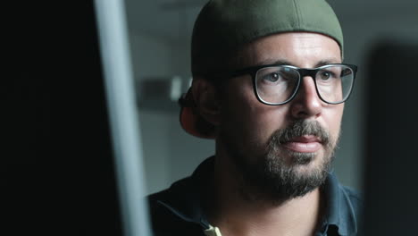 Front-shot-of-Hipster-guy-with-glasses-and-baseball-cap-looking-at-two-computer-screens-concentrated