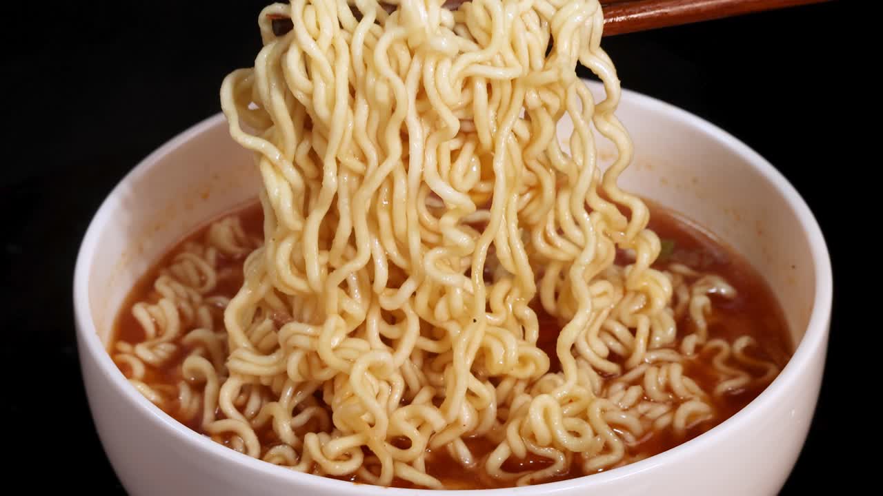 Chopsticks grasp and lift cooked instant noodles from a white bowl filled with broth, under bright studio lighting and a clean black background