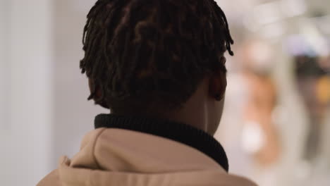 back shot of young black man walking through modern shopping mall corridor wearing denim jacket and hoodie, passing store windows with sale signs and mannequins under bright overhead lighting