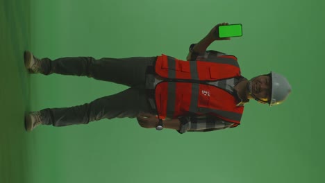 full body of asian male engineer with safety helmet smiling and showing green screen smartphone to the camera while standing in the green screen background studio, during sunset or sunrise time