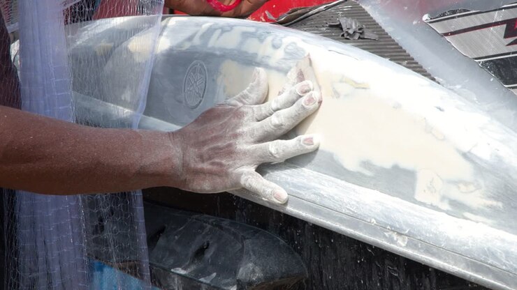 A mechanic uses sandpaper to smooth body filler on a personal watercraft under bright daylight