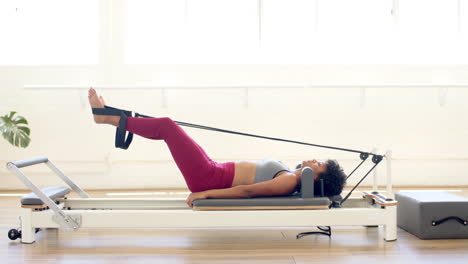 Biracial-woman-wearing-sportswear-exercising-on-Pilates-reformer