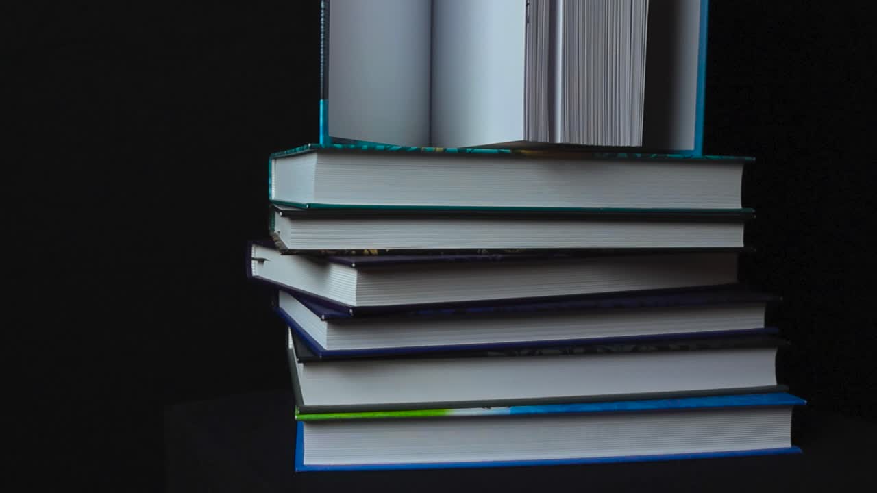 Footage spinning around hardcover thick colorful books that are in a pile on top of each other and the book on top is standing vertically and is opened with white pages visible. On black background.