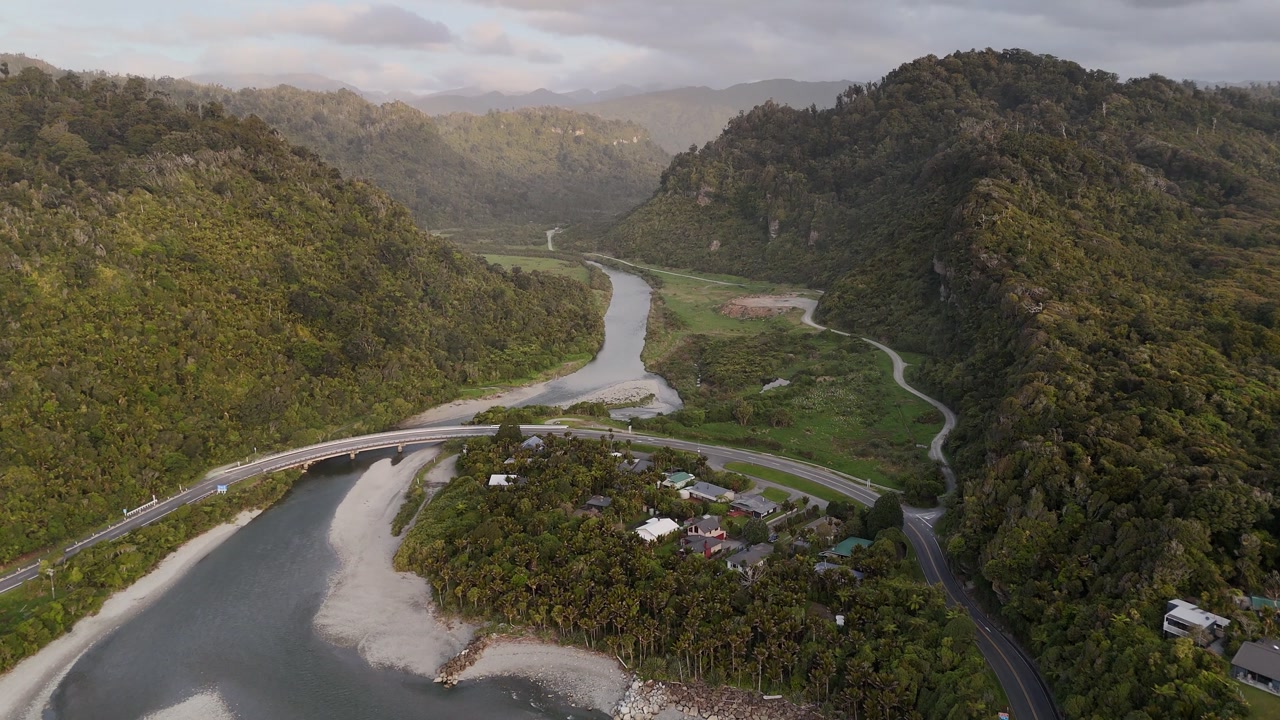 Aerial drone view of Punakaiki River curving through lush forested hills on New Zealand’s South Island West Coast, with bridge crossing and warm sunset light over tranquil landscape