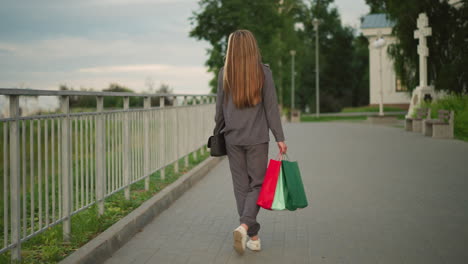 back view of lady in grey clothing walking along iron rail holding shopping bags and black handbag, with blurred view greenery in background, casual outdoor scene during a calm evening stroll