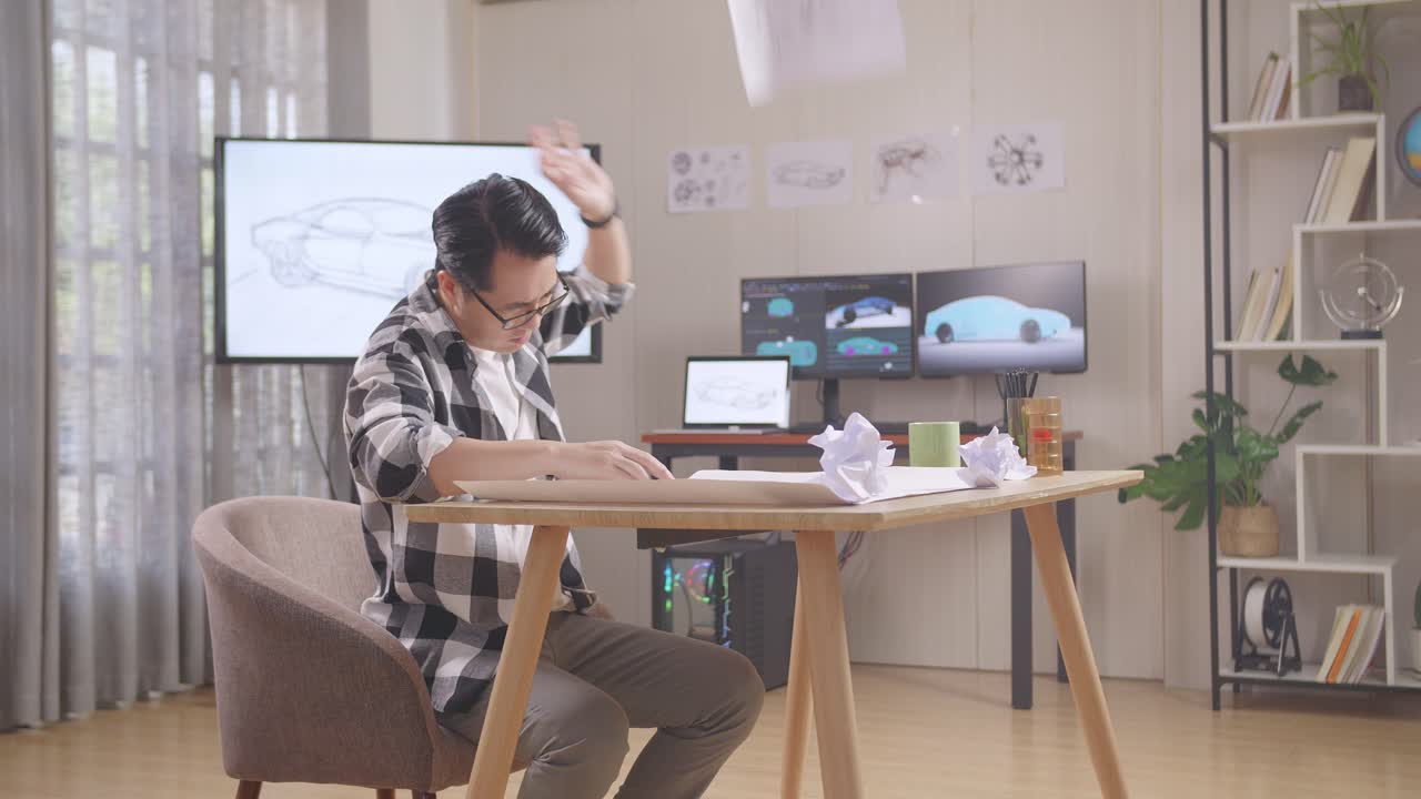 Asian Male Throws Away Papers While Working On A Car Design Sketch On Table In The Studio With Tv And Computers Display 3D Electric Car Model