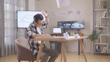 asian male throws away papers while working on a car design sketch on table in the studio with tv and computers display 3d electric car model