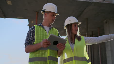 dos ingenieros un hombre y una mujer con cascos blancos con una tableta en el sitio de construcción condenan el plan de construcción del edificio