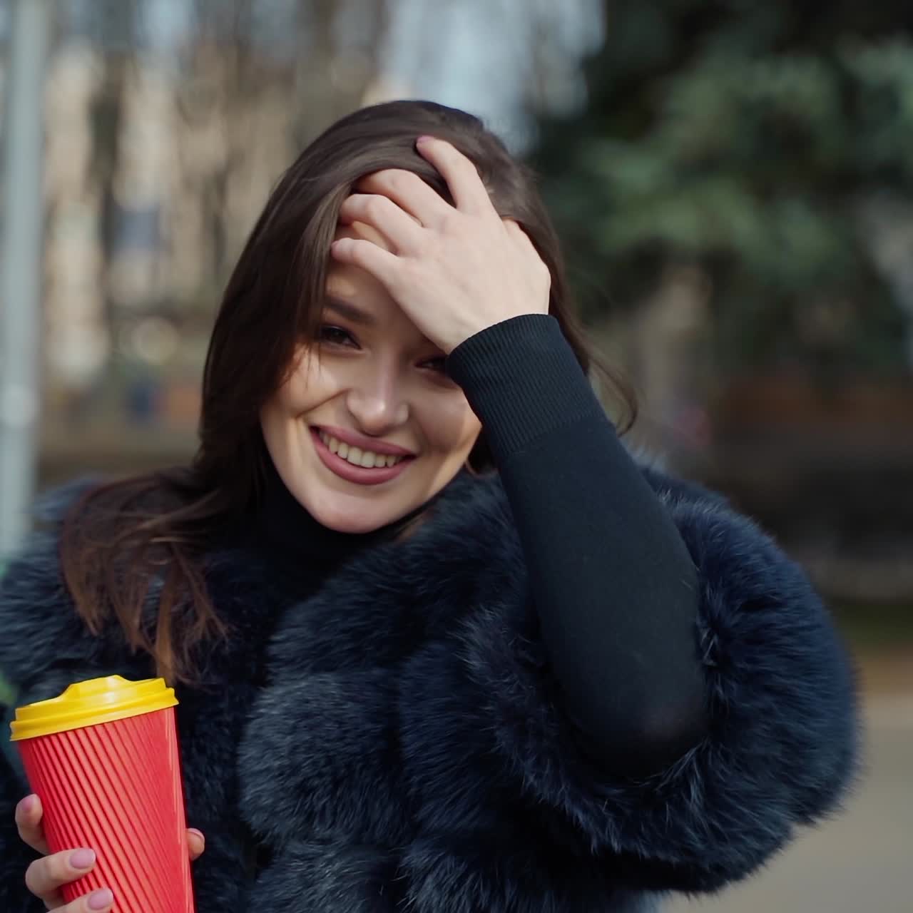 Portrait of a happy girl in a sunny day in winter. Beautiful woman in fur coat holds a cup of hot drink in the park and smiling at camera. Slow motion.