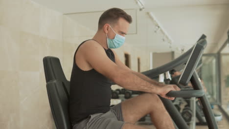 strong male with face mask uses an exercise machine and drinks water in the gym