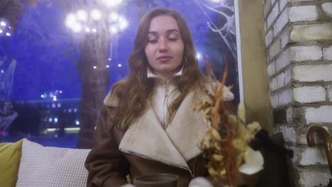 cheerful lady in brown shearling jacket sitting in cozy cafe, resting forward on table with a relaxed and joyful expression, moving cars visible through window in background, warm cafe ambiance