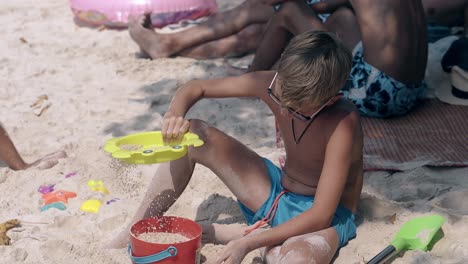 Junge-In-Blauen-Badehosen-Sät-Sand-Durch-Sieb-Am-Strand