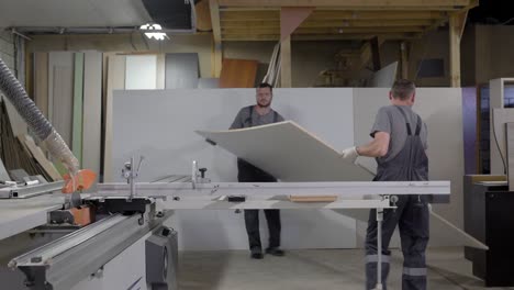dos trabajadores están poniendo tablero de partículas de madera en una máquina de corte en un taller de fabricación de muebles