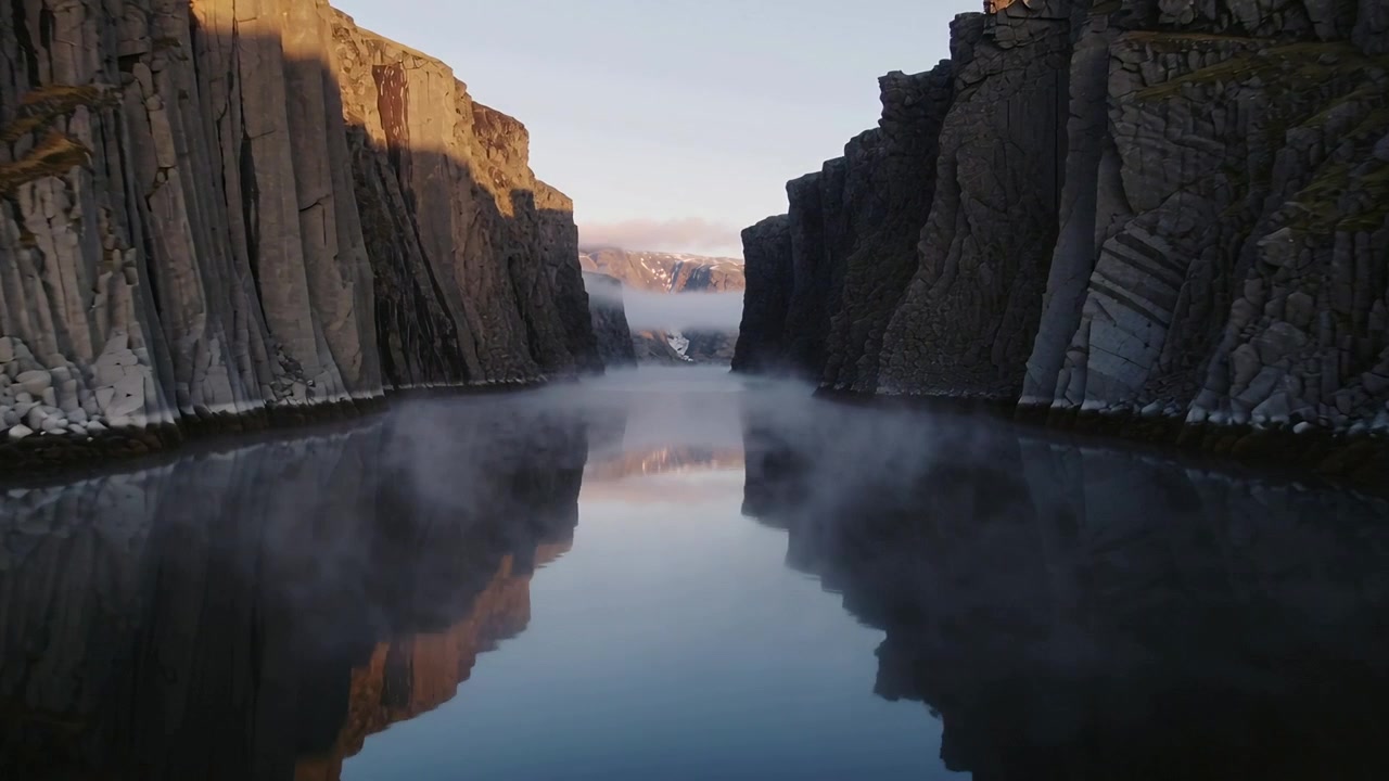 Gliding drone approaching channel in fjord, revealing snowy ridges with mist and cliff reflection