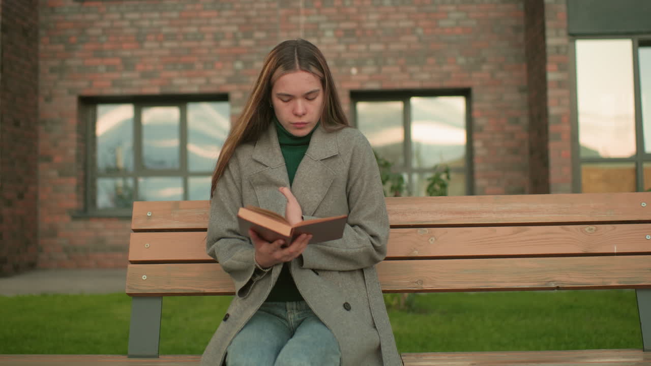 Femme assise sur un banc en bois à l’extérieur, tenant un livre ouvert tout en ajustant son manteau, vêtue d’un pardessus gris et d’un col roulé vert, concentrée sur sa lecture, avec un immeuble résidentiel en brique et de la verdure en arrière-plan.
