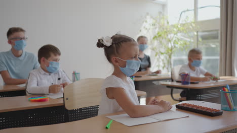 los niños en la escuela se sientan en el aula y levantan las manos respondiendo a las preguntas del maestro en máscaras protectoras en cámara lenta