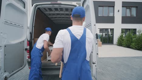 two young workers of removal company unload boxes and furniture from minibus into customer's home