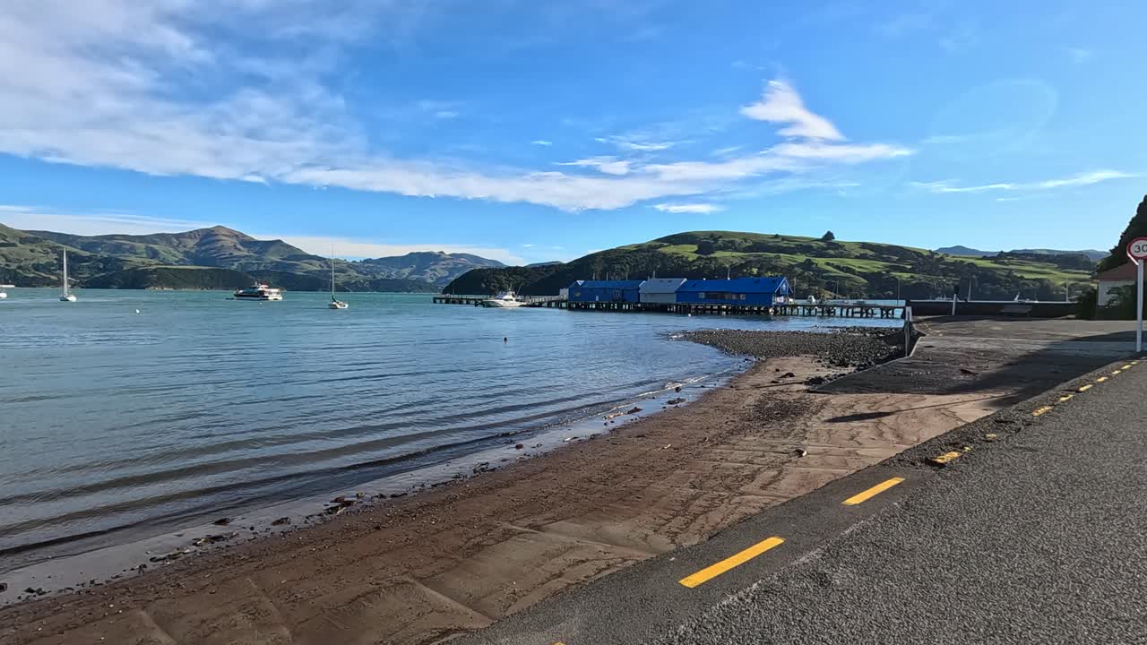 A serene drive along Akaroa's waterfront, capturing calm waters, boats, and vibrant sunrise hues in a tranquil setting