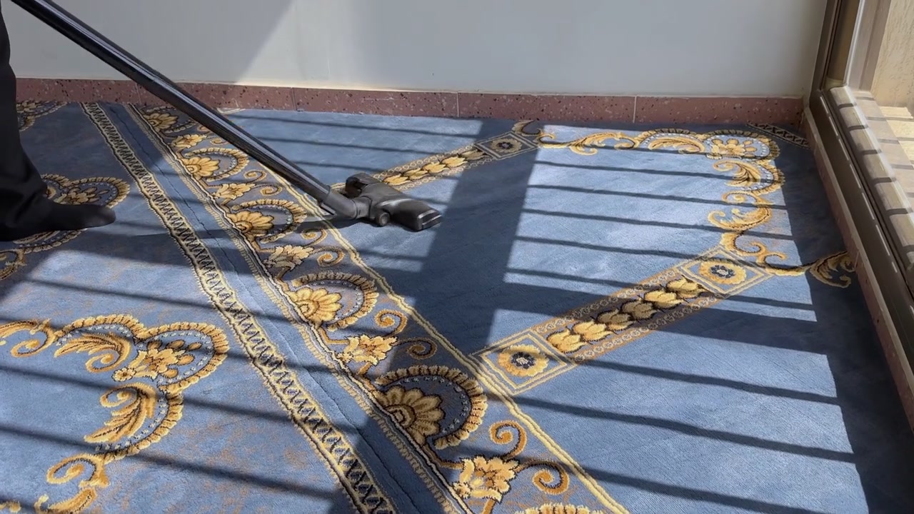 Una persona con calzini neri passa l'aspirapolvere su un tappeto a motivi blu e oro all'interno di una stanza illuminata dal sole. La forte luce naturale del giorno proietta ombre lineari delle finestre sul pavimento durante la manutenzione interna quotidiana.