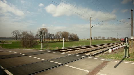 Eine-Rote-Elektrolokomotive-Mit-Einem-Markanten-Weißen-Streifen-Fährt-Schnell-An-Einem-Bahnübergang-Im-Europäischen-Stil-Vorbei,-Vor-Dem-Hintergrund-Einer-üppigen-Grünen-Wiese-Und-Eines-Blauen-Himmels