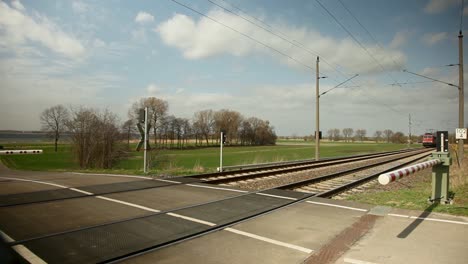 red electric locomotive, with a distinctive white stripe, moves swiftly past a european style railway crossing amidst the backdrop of a lush green field and blue sky